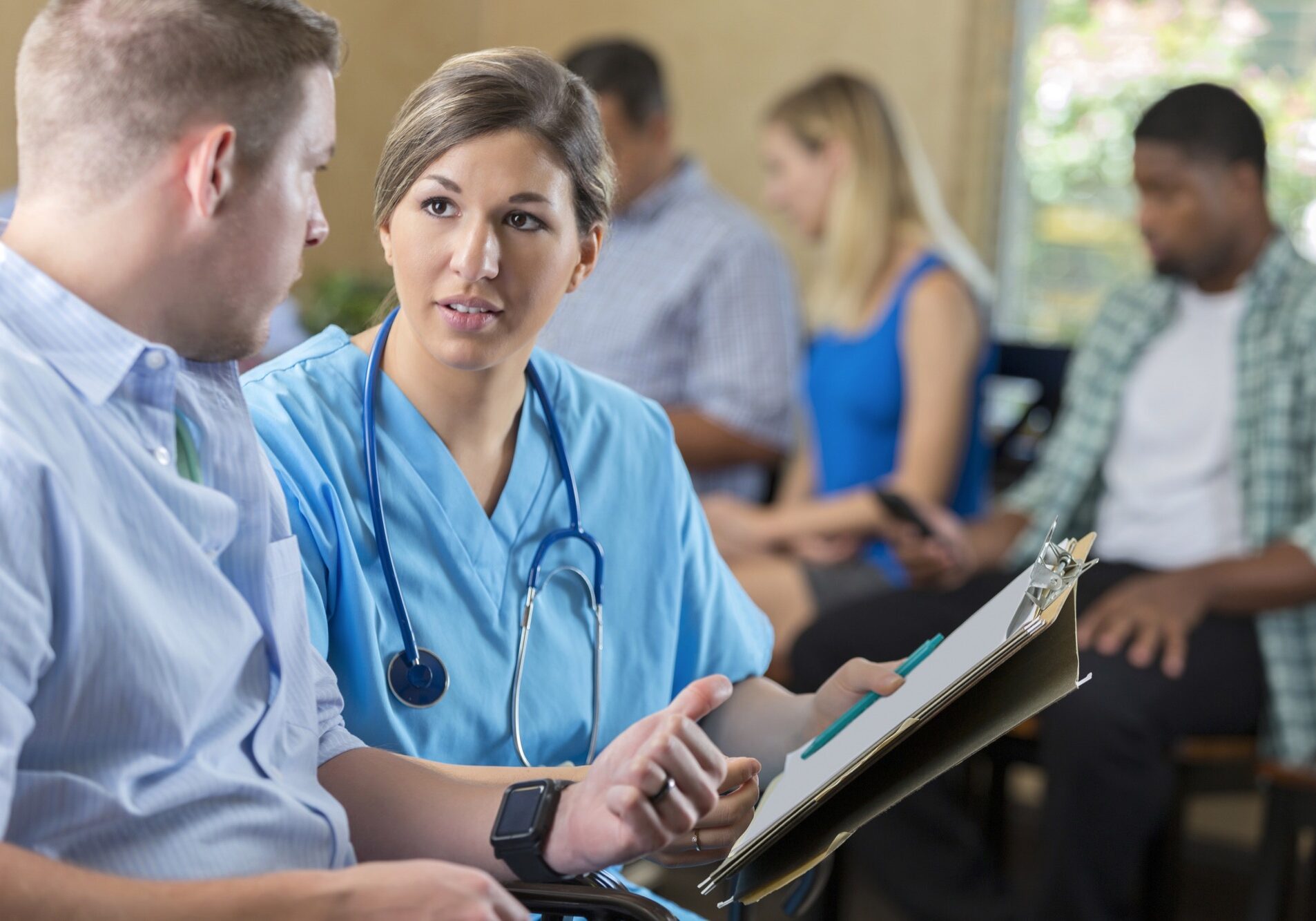 Mid adult Caucasian woman is nurse in hospital emergency room triage center. She is examining and talking with a patient, while other patient's wait behind them in waiting room. Nurse is reviewing paperwork with male patient.