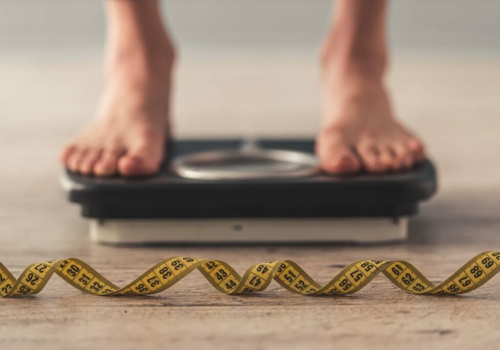 Cropped image of woman feet standing on weigh scales, on gray background. A tape measure in the foreground